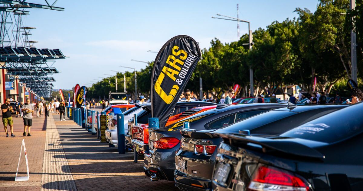 A row of luxury and sports cars lined up at a "Cars & Culture" outdoor auto show with spectators walking along Olympic Boulevard.