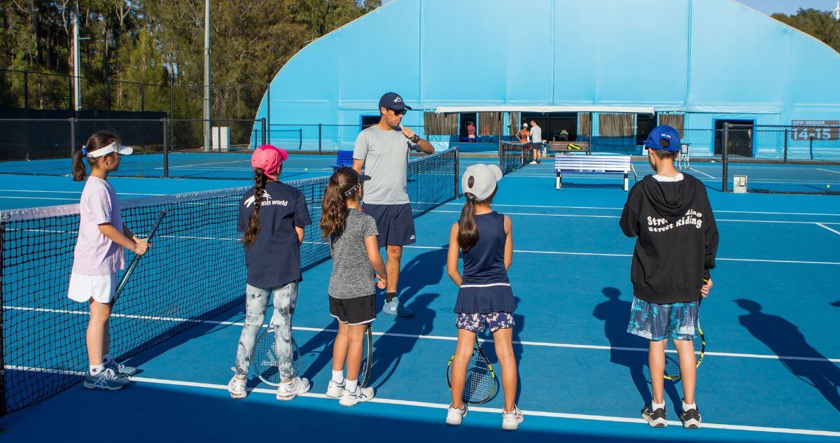 A tennis coach instructs a group of young children on a bright blue outdoor tennis court.