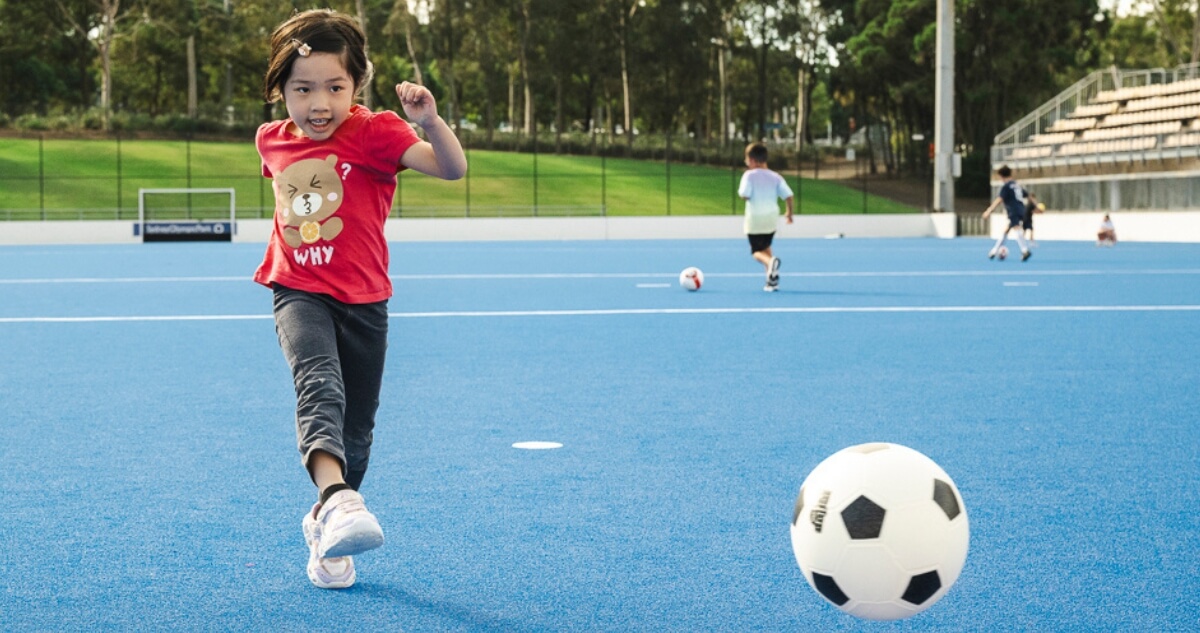 A child in a red shirt plays soccer on a blue field with other children and a goalpost in the background.