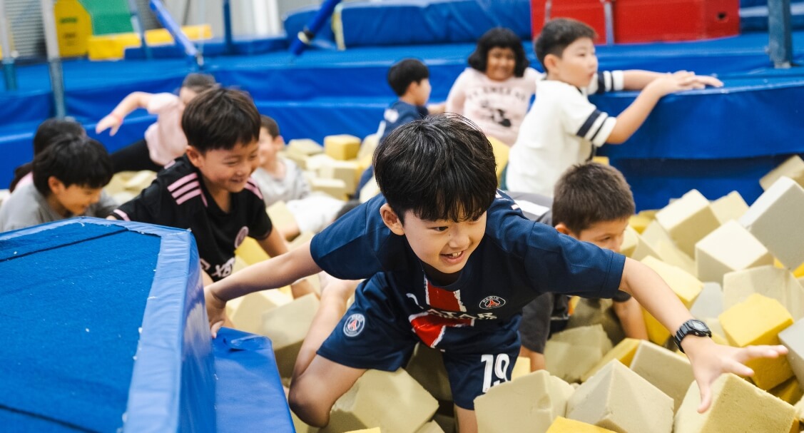 Children play and climb across soft foam blocks in an indoor gymnastics area with blue padded equipment and colourful mats.