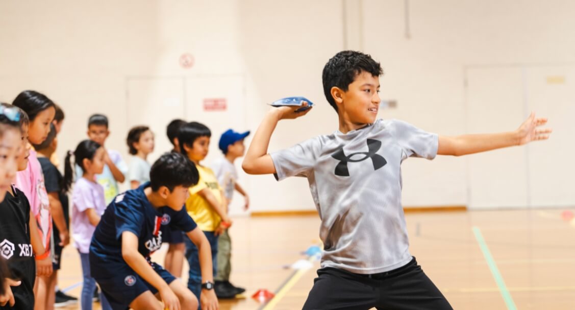 A child in a gymnasium is mid-throw with a small blue bean bag while a line of other children in sportswear watch and wait their turn during an organised indoor activity.