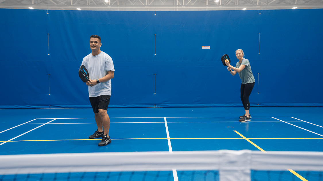 Two people play pickleball on an indoor court, standing ready on opposite sides of the net against a bright blue backdrop.