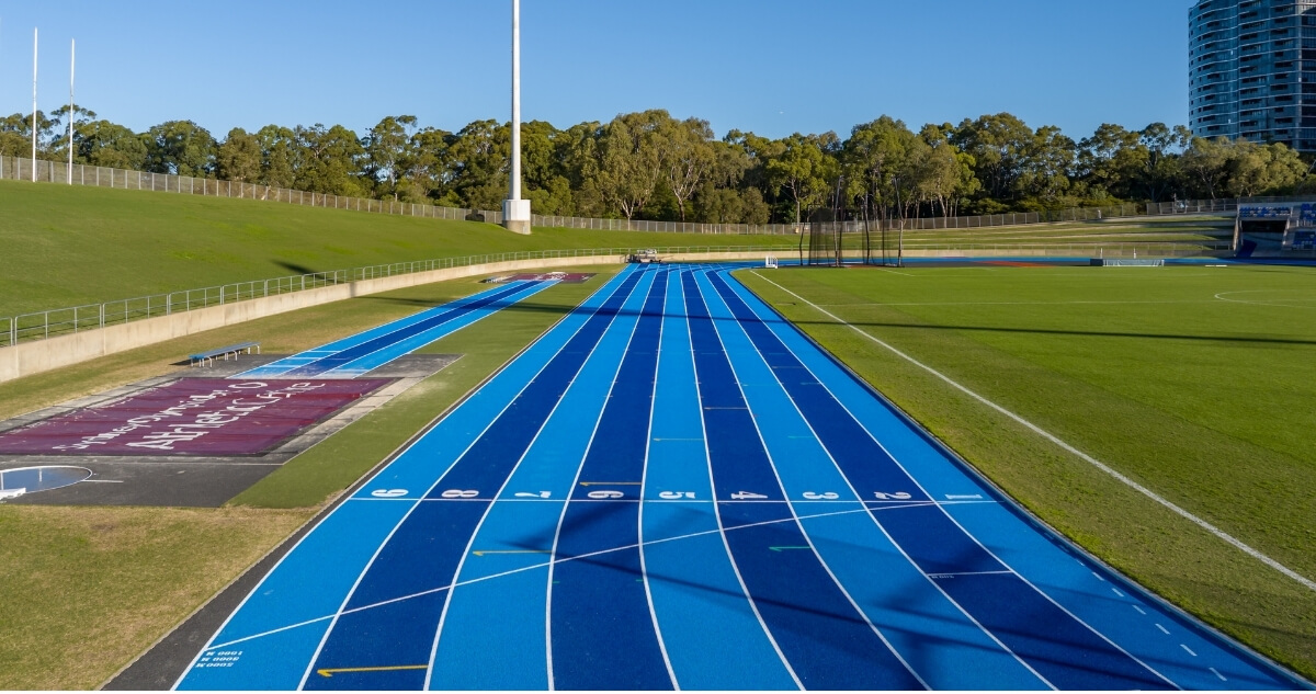 Blue athletics running track with numbered lanes beside a green sports field, set within Sydney Olympic Park Athletic Centre under a clear blue sky.