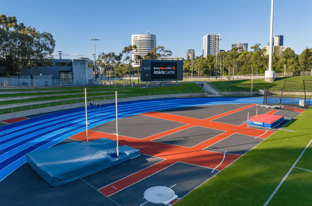Outdoor athletics track and field facility with blue running lanes, pole vault and throwing areas, and a large screen 