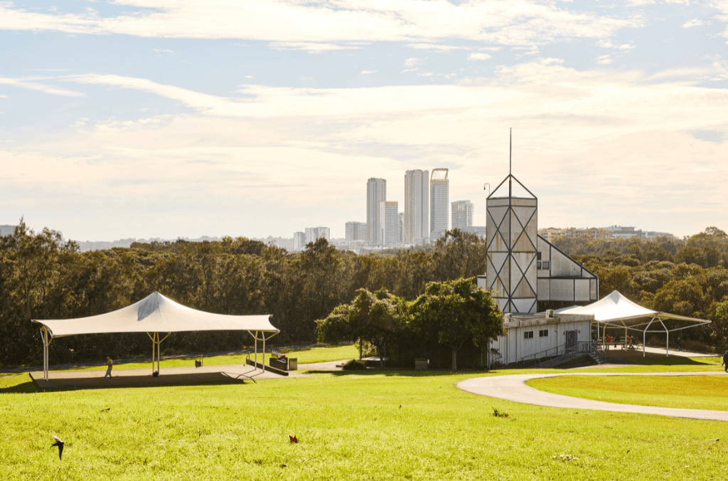 A sunlit park features shade sail shelters, a small building, and a winding path on a grassy hill, with a distant city skyline rising beyond the trees.